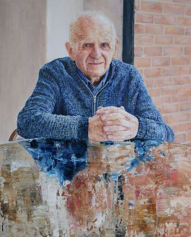A older man with grey hair and wrinkles sits at a glass table, he is wearing a blue zip up cardigan and matching shirt in a house next to a back door.