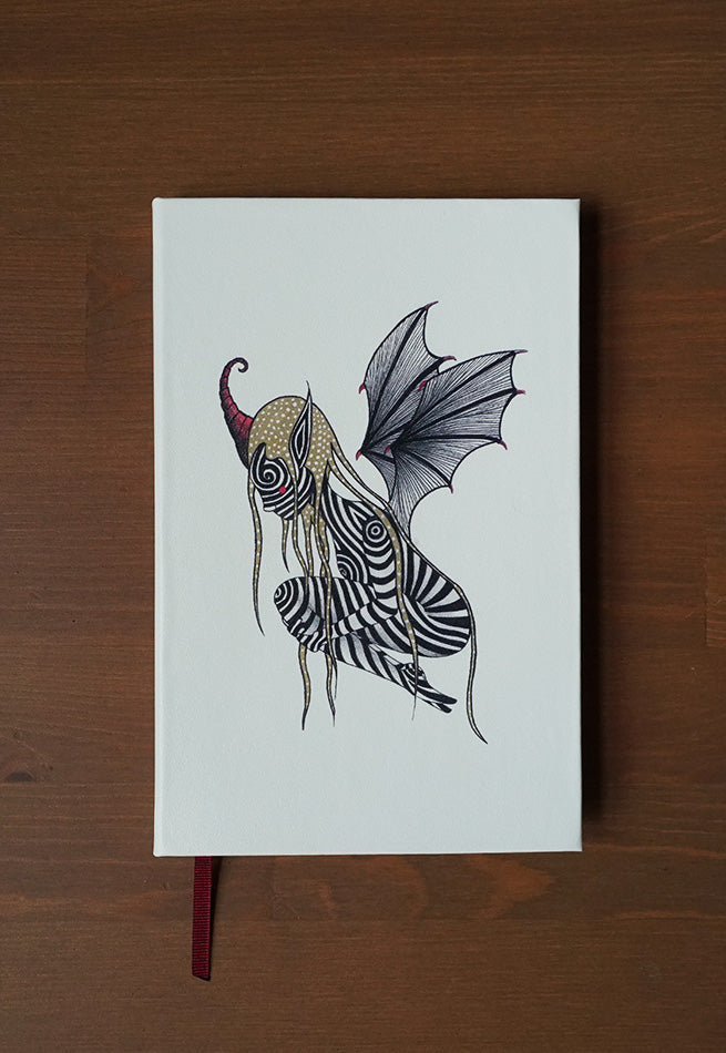 An ivory coloured hardback notebook with a design on the cover. A black and white striped elf with wings and a horn. Colours of the illustration include black, white, red and gold.